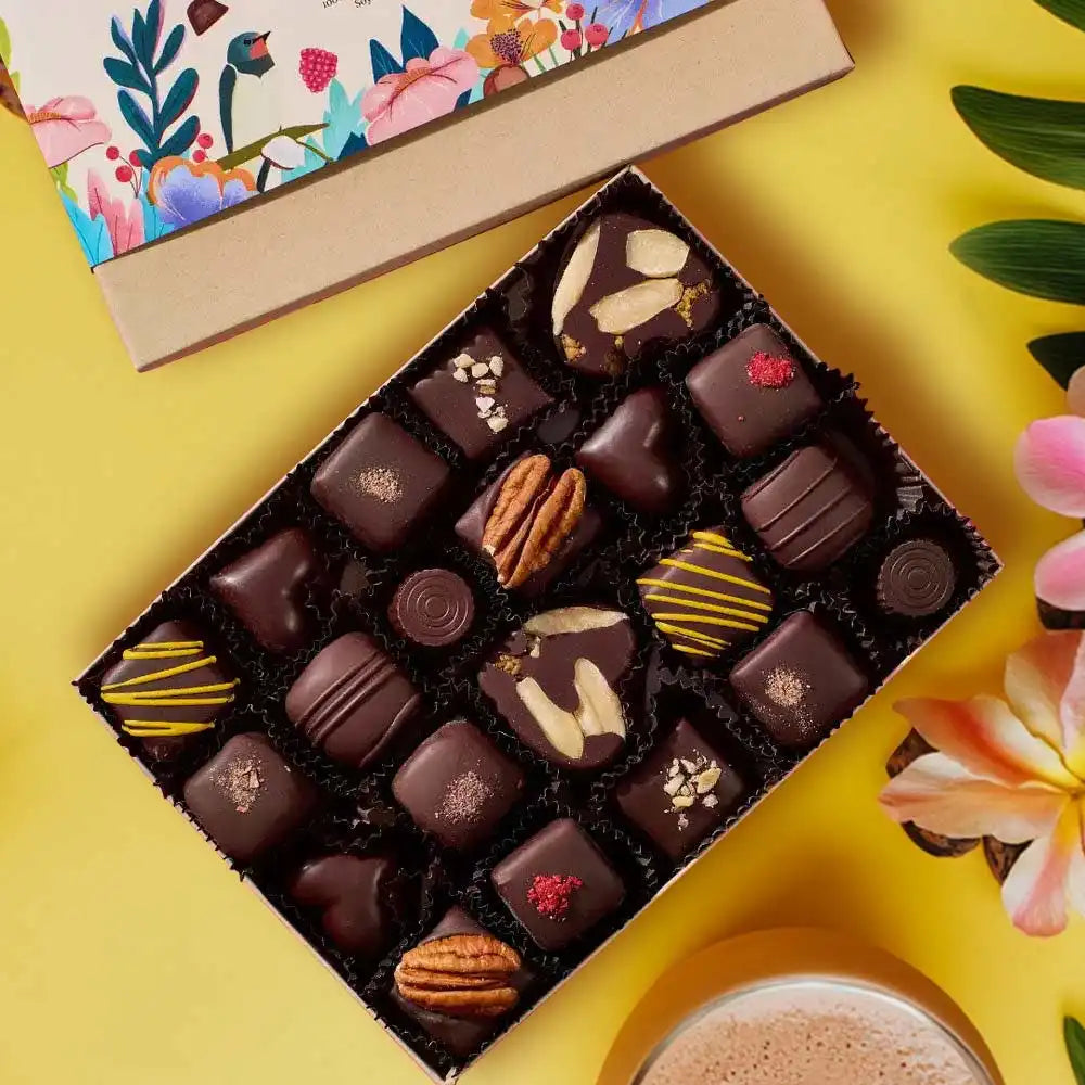 Assorted chocolates in a box on a yellow surface with flowers and a colorful box in the background